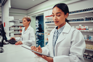 Two female pharmacists, one using a computer, one using a tablet